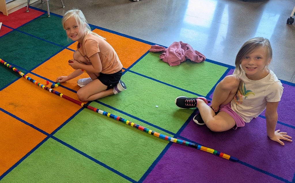 Two Kindergarten students sitting on a colorful checkered classroom rug next to an extra-long line of connected plastic counting cubes.