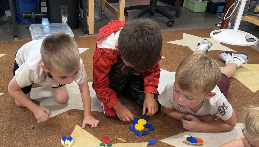 Three Kindergarten students leaning over a carpet as they collaborate to create shapes and designs using colorful plastic pattern blocks.