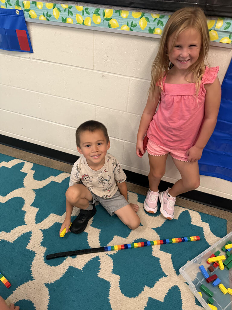 Two smiling Kindergarten students posing on a teal and white pattern rug with a long line of connected colorful Unifix cubes.