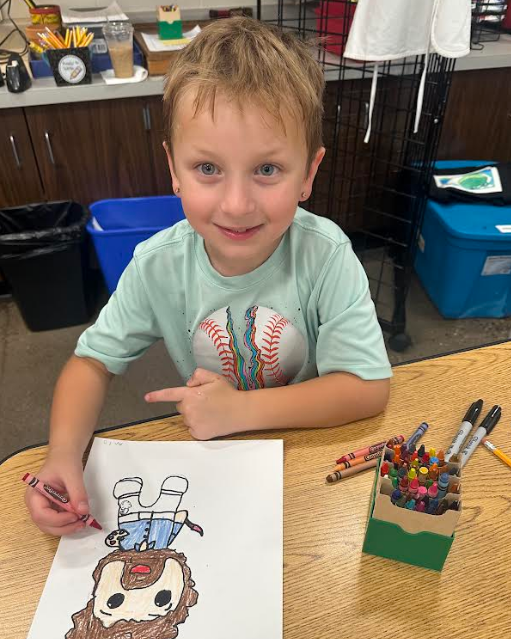 Young student pointing to his completed Bob Ross coloring page at a desk.