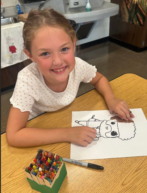 Young student smiling at a desk while coloring a Bob Ross drawing with crayons.