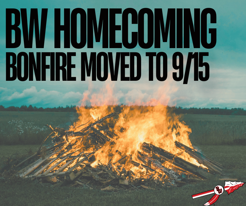 Large, blazing bonfire burning outdoors in an open field against a cloudy sky. Bold black text at the top reads "BW HOMECOMING BONFIRE MOVED TO 9/15". The Baldwin-Woodville spear and feather logo appears in the bottom right corner.