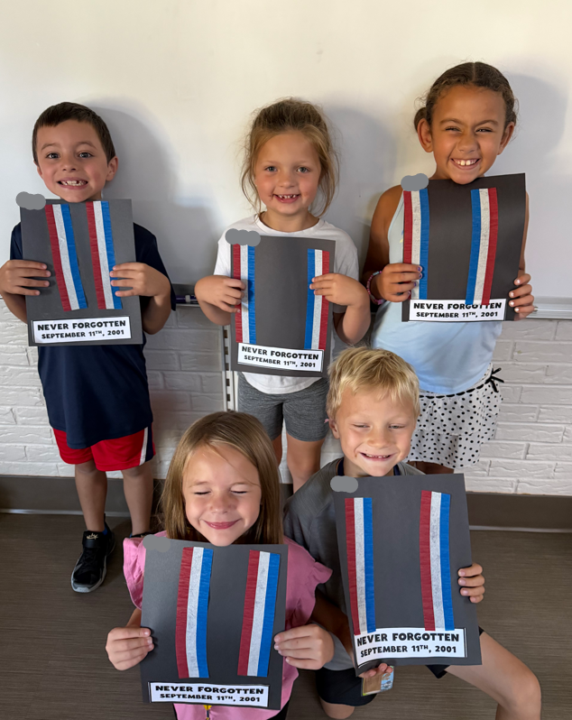 Five students smiling in a classroom, holding up construction paper Patriot Day crafts depicting the Twin Towers lined with red, white, and blue patriotic ribbons and a banner that reads "NEVER FORGOTTEN SEPTEMBER 11TH, 2001."