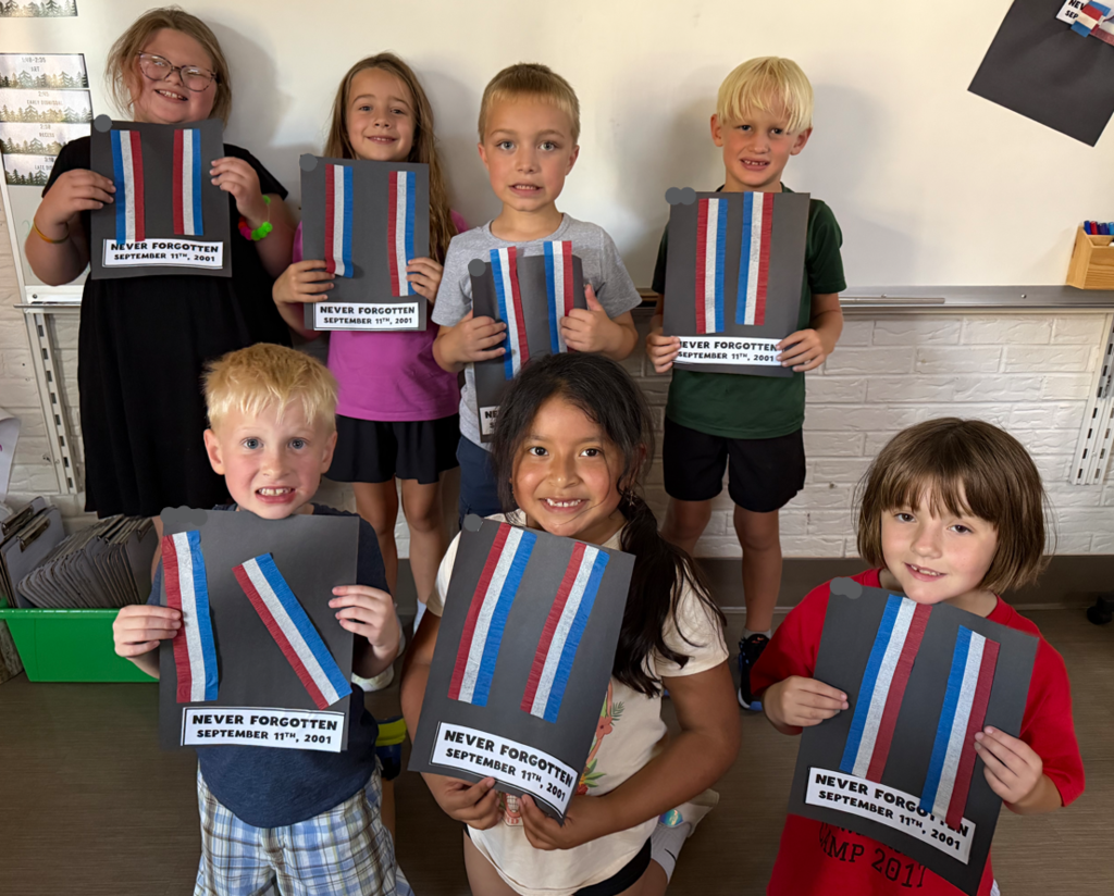 A group of seven elementary students proudly posing in a classroom while displaying their handmade September 11th commemorative art crafts on dark paper.