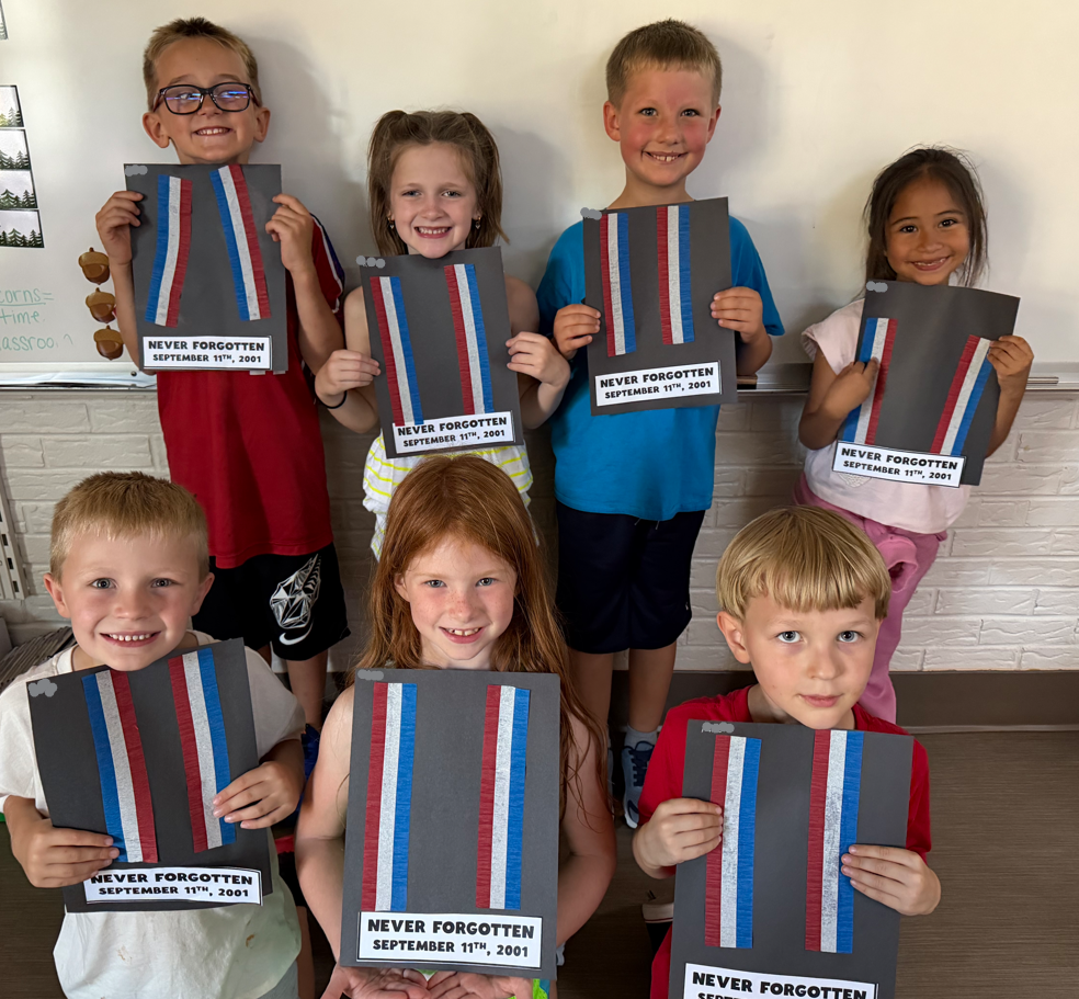 Seven young students smiling in a classroom setting, holding up black paper craft projects featuring red, white, and blue paper stripes representing the Twin Towers with a printed sign reading "NEVER FORGOTTEN SEPTEMBER 11TH, 2001" attached at the bottom.