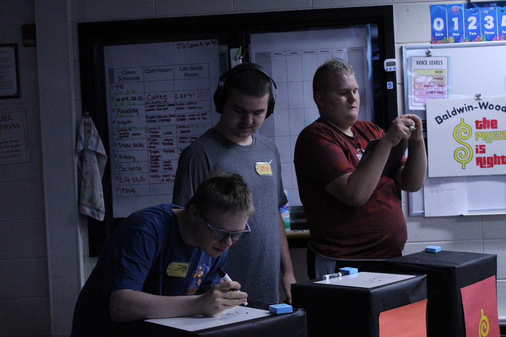 Three students working at their game podiums, writing price estimates on dry-erase boards during a classroom activity.