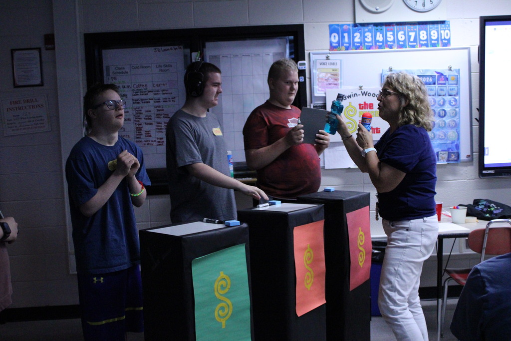 A teacher showing items to three students standing behind game podiums during a classroom "The Price is Right" activity at Baldwin-Woodville.