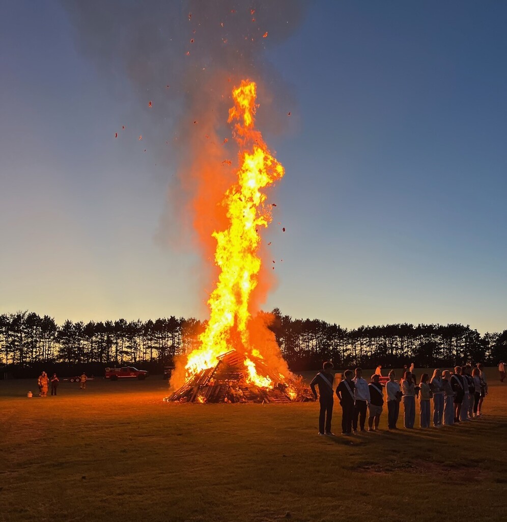 A towering, bright bonfire constructed from wooden pallets burning brightly in a grassy field at dusk, with high flames shooting into the evening sky. Members of the homecoming court stand in a line on the right side, watching the fire.