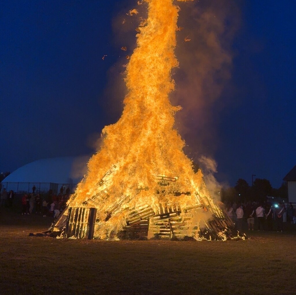 A close-up view of a massive bonfire made of stacked wooden pallets, engulfed in roaring orange and yellow flames against a dark blue night sky.