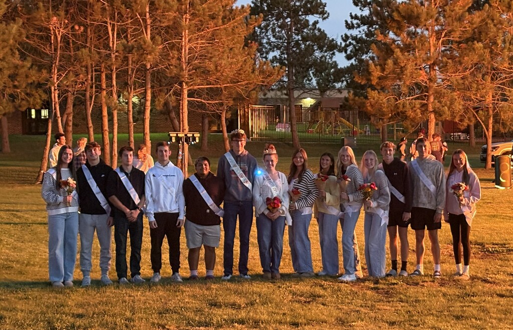 The 2026 Homecoming Court and class representatives standing in a long line on a grassy field at sunset, wearing sashes and holding flower bouquets, with pine trees in the background illuminated by warm golden light.