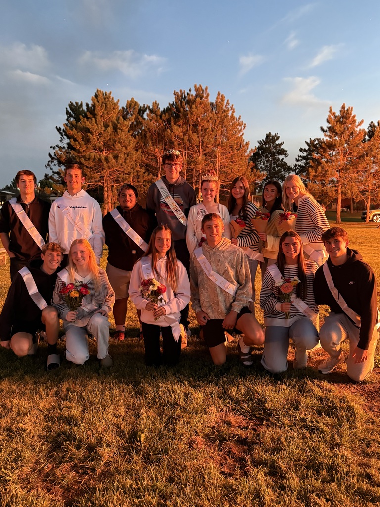 A group photo of the Homecoming Court illuminated by the warm glow of the bonfire light. The Homecoming King and Queen wear crowns in the center of the back row, surrounded by fellow court members kneeling and standing together outdoors.