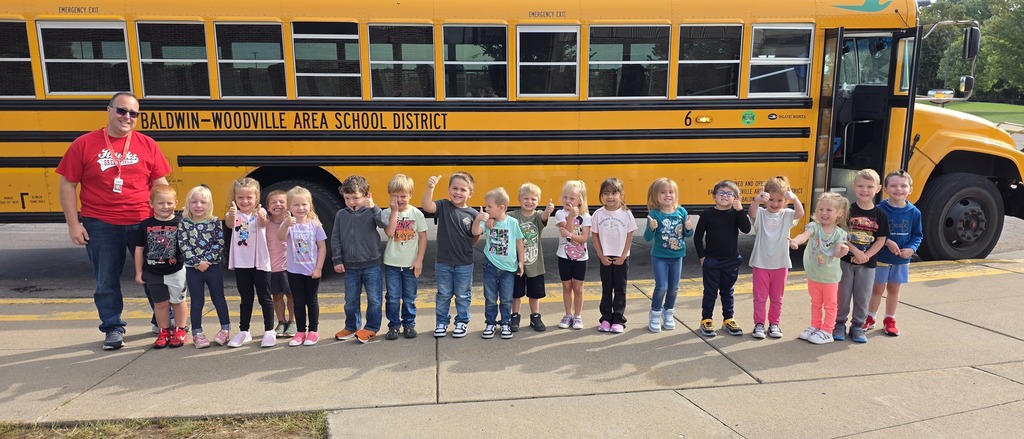 group of young students and a staff member wearing a red shirt stand in a line along the sidewalk, giving thumbs-up gestures for a photo in front of a yellow Baldwin-Woodville school bus.