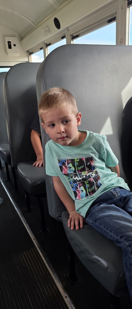 A young student in a light green graphic t-shirt and jeans sits attentively in a high-backed seat inside a school bus.