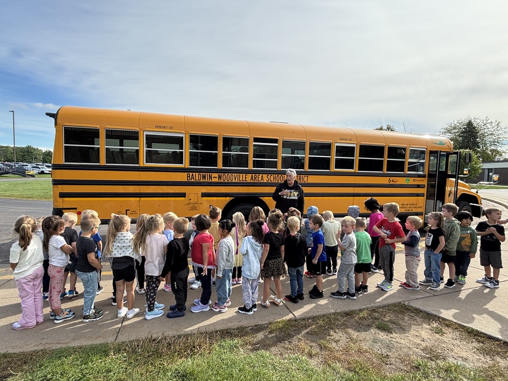 A large group of young students lines up along the sidewalk beside a yellow Baldwin-Woodville Area School District bus, listening attentively to bus driver Faye during a bus safety lesson.