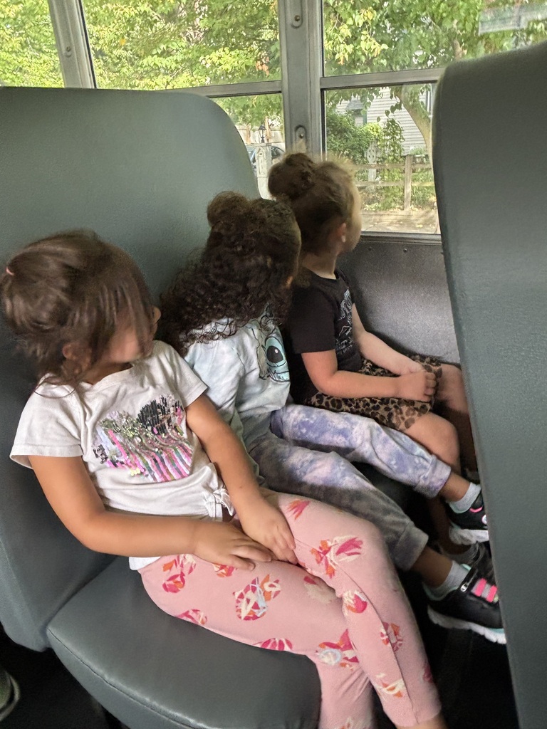 Three young students sit together on a high-backed vinyl seat inside a yellow school bus, looking out the window during a bus safety lesson.