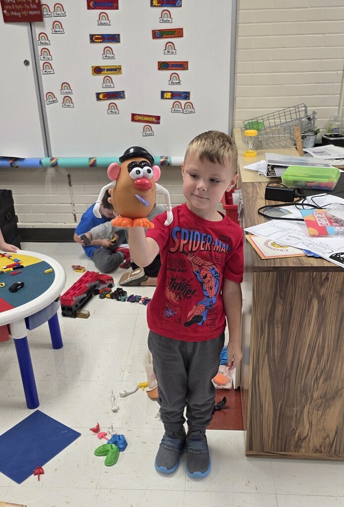 A young student in a red Spider-Man shirt proudly holding up a completed Mr. Potato Head toy in a 4K classroom.
