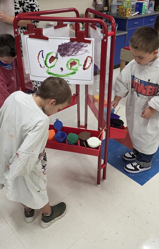 Two young students wearing white paint smocks painting creative artwork at a red double-sided easel in the classroom.