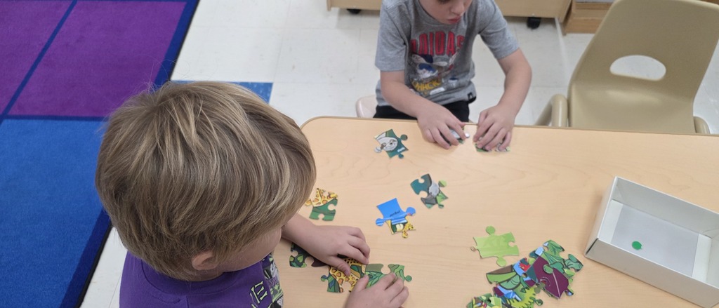 Two young 4K students sitting at a light wood table collaborating and putting together colourful floor puzzles.