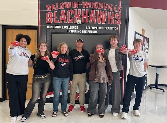 Six Baldwin-Woodville High School students and Mr. Dachel posing together in front of a Baldwin-Woodville Blackhawks wall graphic, with each student holding up a small red shoe award.