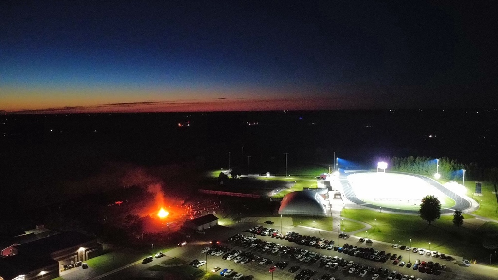 Wide aerial view of the Baldwin-Woodville school campus at dusk, capturing the glowing horizon, illuminated football stadium, parking area, and the bright bonfire.