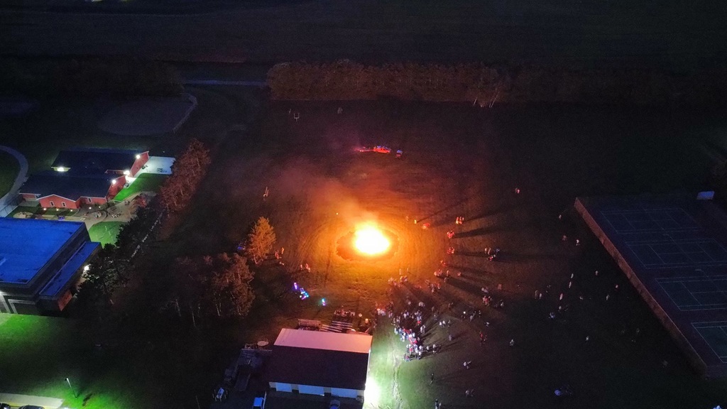 Nighttime aerial drone photo showing a glowing orange bonfire on the school grounds with students and community members gathered around it in a large circle.
