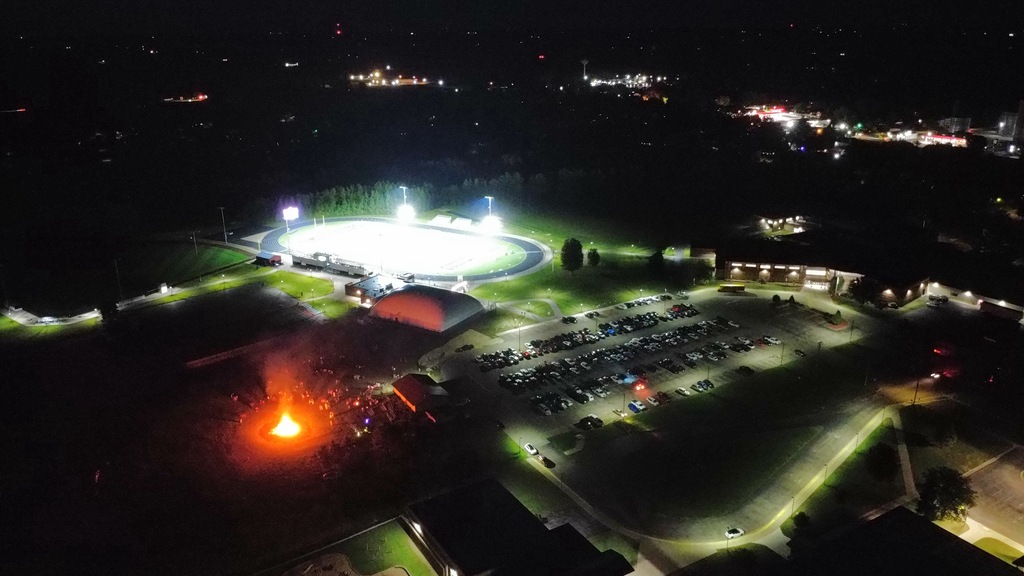 High-angle drone view of the school campus at night, showing the brightly lit football field, a packed parking lot, and the glowing bonfire nearby.