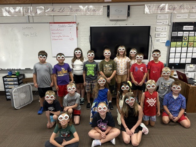 A large group of elementary school students standing and sitting in a classroom, posing for a photo while wearing paper craft "scuba goggles" over their eyes. A large white display board and TV monitor sit in the background.