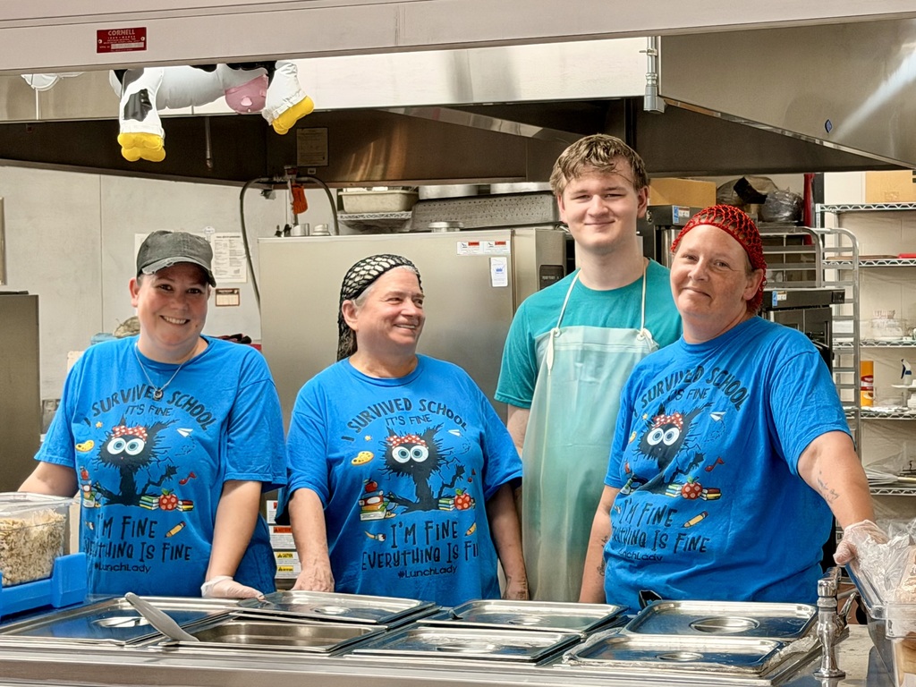Four high school cafeteria staff members standing together behind a stainless steel serving counter in a kitchen. Three women on the left and right wear matching bright blue t-shirts that say "I Survived School... I'm Fine Everything is Fine" with a humorous cartoon monster graphic, while a young man in a teal shirt and white apron stands among them, all smiling for the camera during Homecoming Spirit Week Twin Day.
