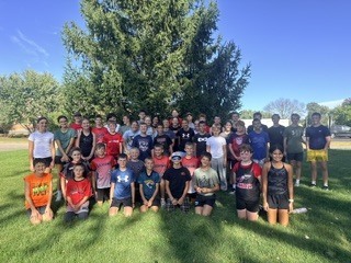 Large group photo of middle school and high school cross country runners smiling together while posing outdoors on a sunny day in front of a tall pine tree.