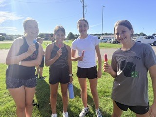 Four female cross country athletes standing outside on the grass, holding popsicles and smiling at the camera after practice.