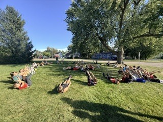 Wide shot of cross country runners lying on the grass in a circle on a sunny day doing abdominal cooldown stretches together.