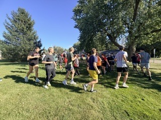 Cross country runners participating in a dynamic warmup activity, jogging together on a grassy field outdoors under clear blue skies.