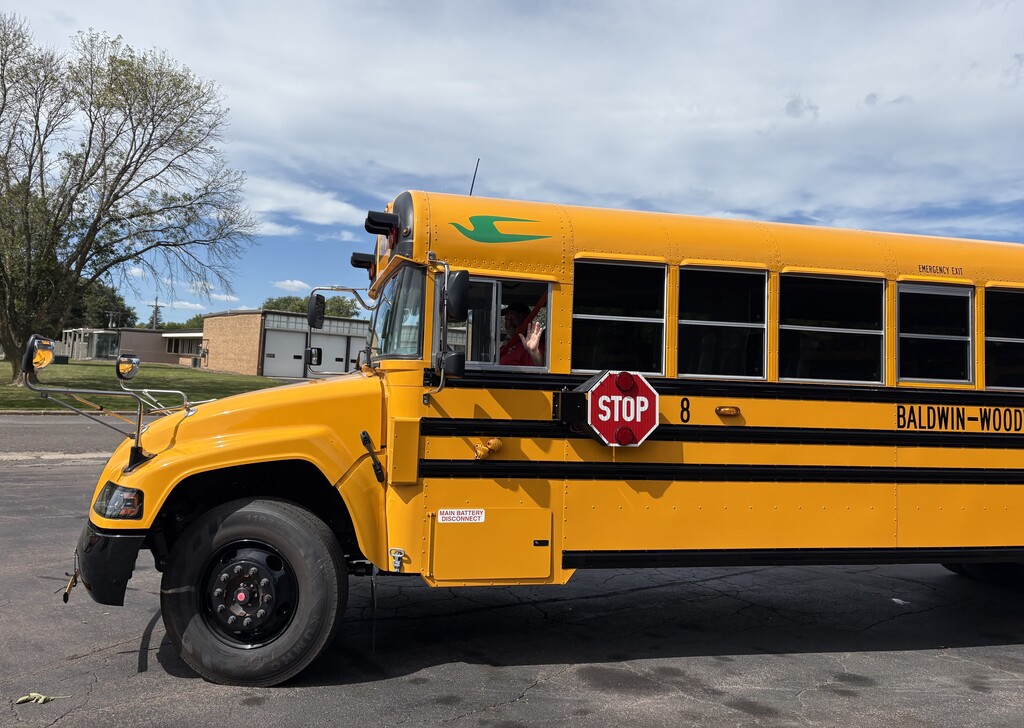 A yellow Baldwin-Woodville Area School District school bus with a green bird emblem on the roof peak, driven by Bus Driver Brad, who is smiling and waving from the driver's side window.