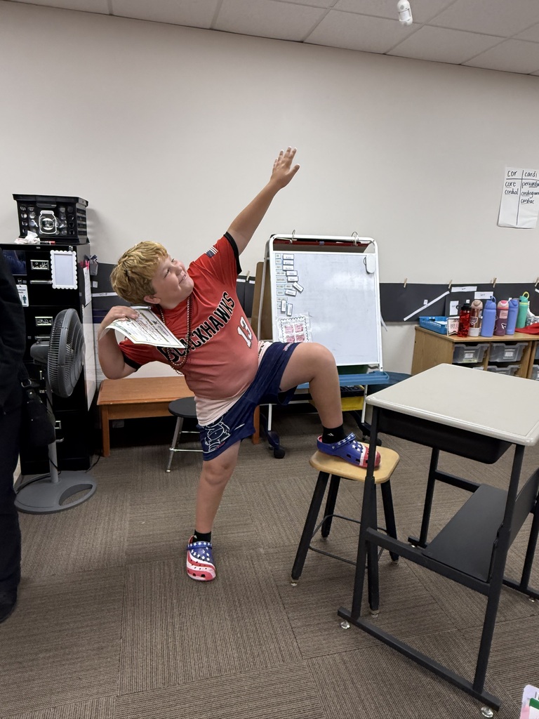 A young boy in a red shirt strikes a playful Heisman trophy-style football pose with one foot up on a stool inside a classroom.