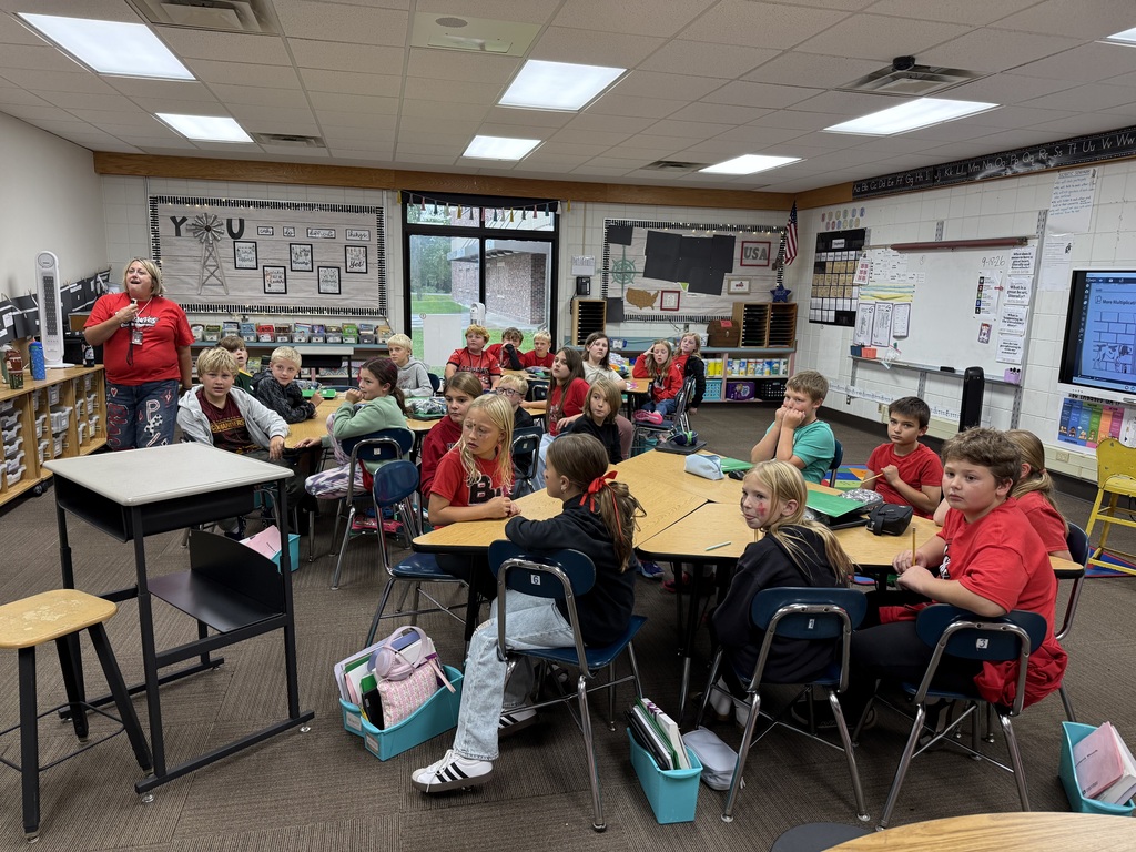 A classroom full of elementary students sitting at their desks, listening attentively while a teacher stands near the front of the room speaking.