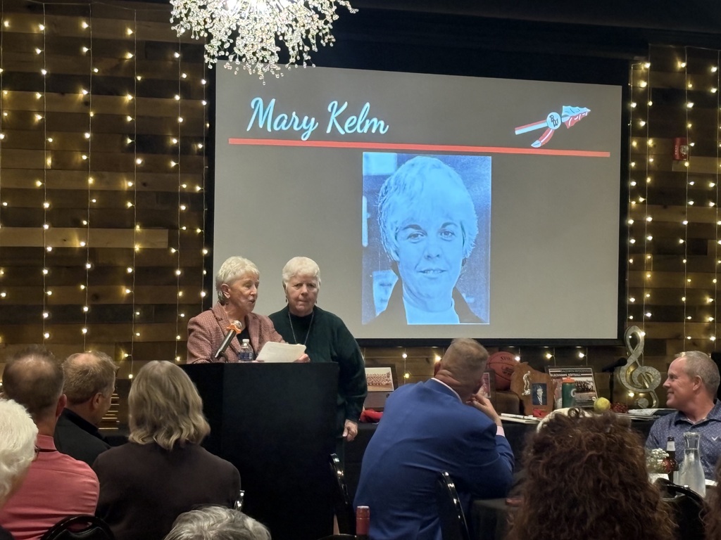 Two women speaking at a podium during a ceremony with a slide reading "Mary Kelm" projected on the screen behind them.