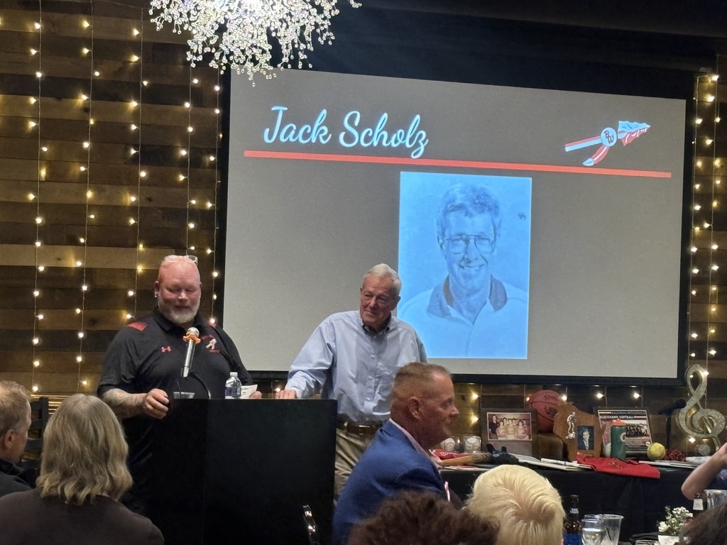 A speaker at a podium addressing the audience alongside another man during an induction ceremony with a slide reading "Jack Scholz."