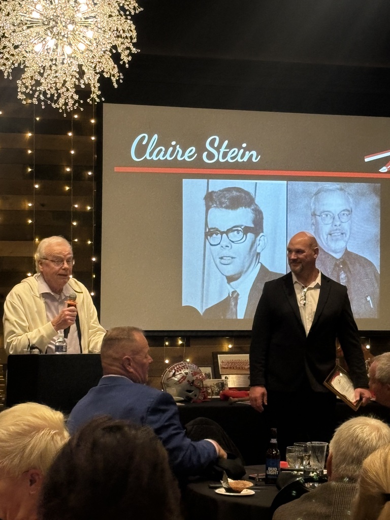 A speaker holding a microphone at a podium next to a man in a suit during a presentation with a slide reading "Claire Stein."