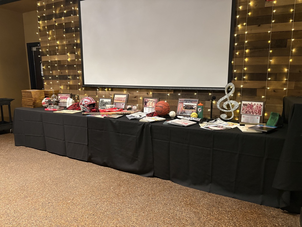 A display table draped in black fabric featuring sports memorabilia, photos, awards, string lights, and a treble clef sculpture under a projection screen.