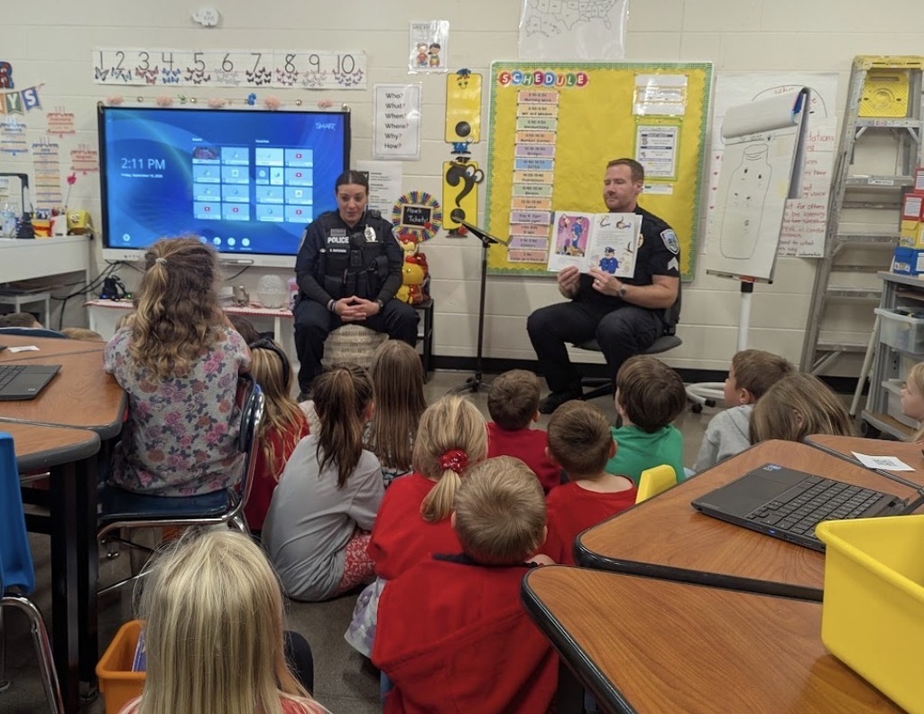 Wide view of a kindergarten classroom where two uniform police officers sit in front of a group of children seated on the floor, holding up a picture book as they read aloud.