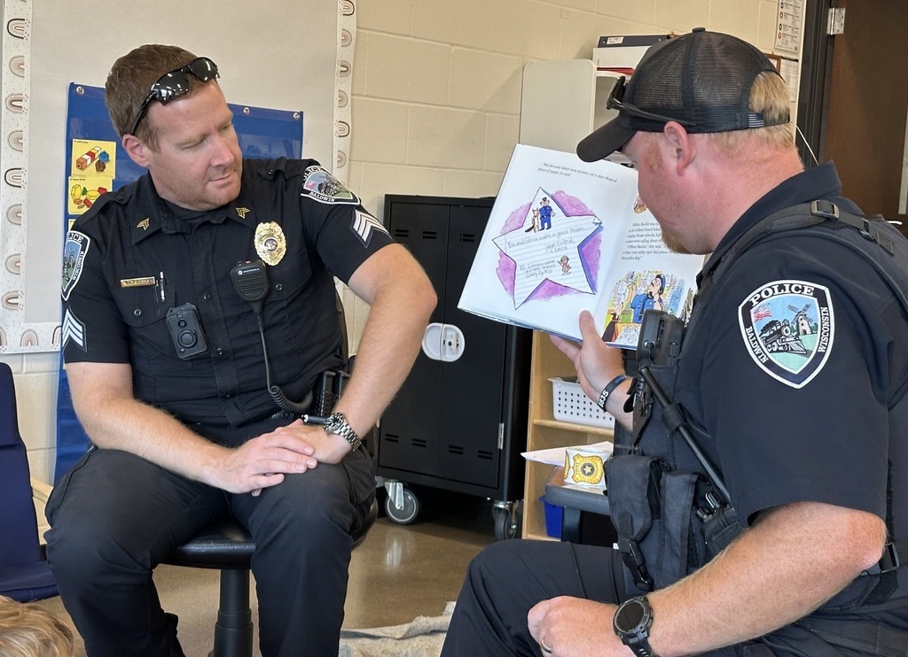 Close-up of two police officers sitting side-by-side in a classroom, with one holding open a picture book displaying a star-shaped drawing while the other officer looks at the page.
