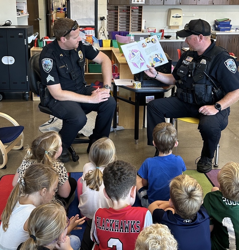 Two police officers in black uniforms sit in chairs in a kindergarten classroom, reading an open picture book to a group of young children sitting on a colorful rug.