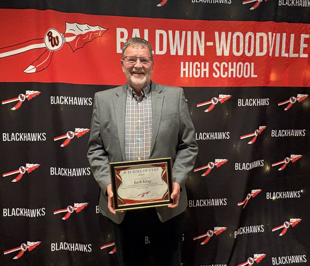 Jeff King smiling and holding an framed Baldwin-Woodville Hall of Fame Class of 2026 award plaque for his late father, Jack King, in front of a red and black Baldwin-Woodville High School Blackhawks backdrop.