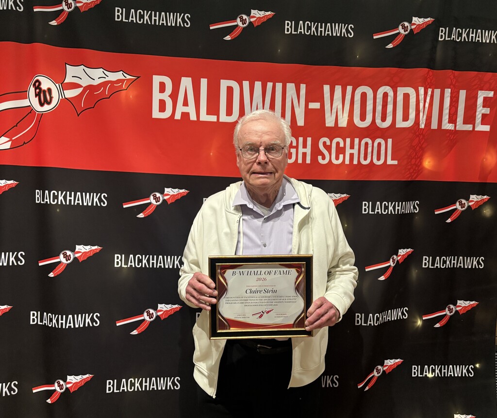Claire Stein smiling and wearing glasses while holding a framed Baldwin-Woodville Hall of Fame Class of 2026 award plaque in front of a red and black Baldwin-Woodville High School Blackhawks backdrop.