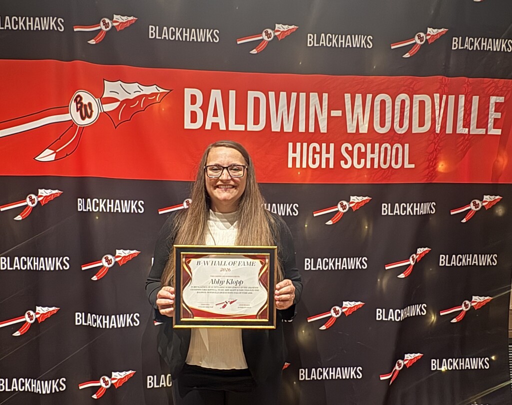 Abby Klopp smiling while holding her framed 2026 Baldwin-Woodville Hall of Fame certificate in front of a red, black, and white backdrop featuring the Baldwin-Woodville High School name and the district spear logo.