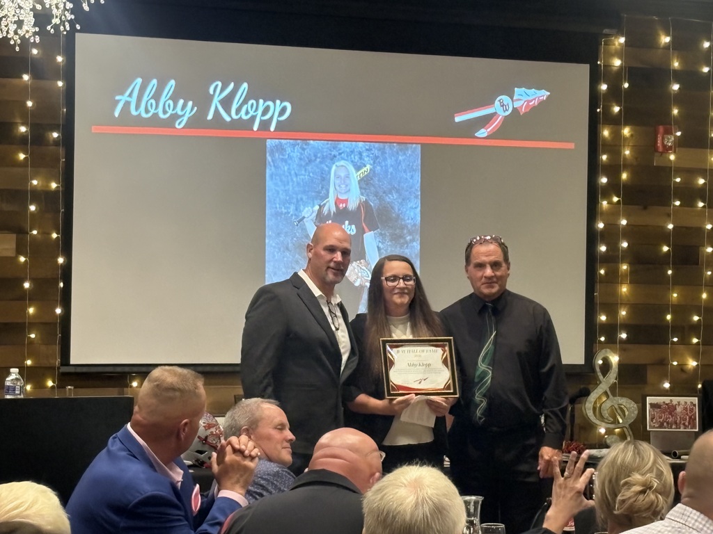 Abby Klopp stands on stage holding her framed Baldwin-Woodville Hall of Fame certificate, flanked by two men in formal attire. In the background, a projection screen displays her name "Abby Klopp," the district spear logo, and her black-and-white high school softball photo.