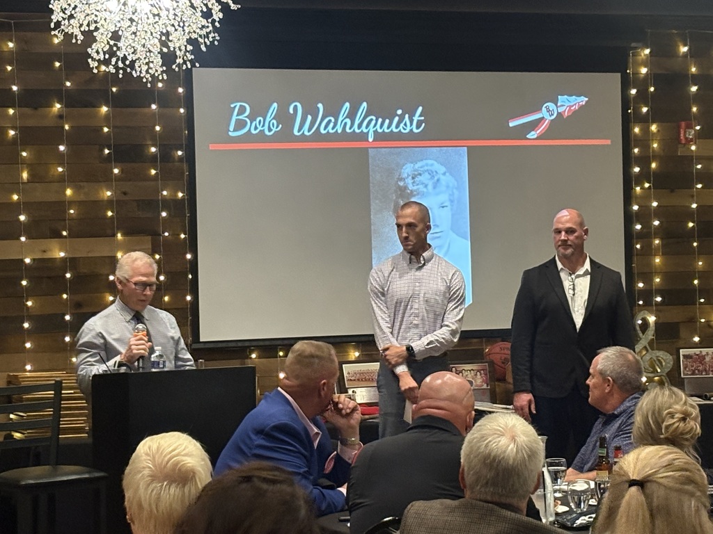 Bob Wahlquist speaking at a podium during the Hall of Fame ceremony. In the background, a large screen displays his name "Bob Wahlquist," his high school photo, and the district spear logo, with two presenters standing on stage beside the screen.