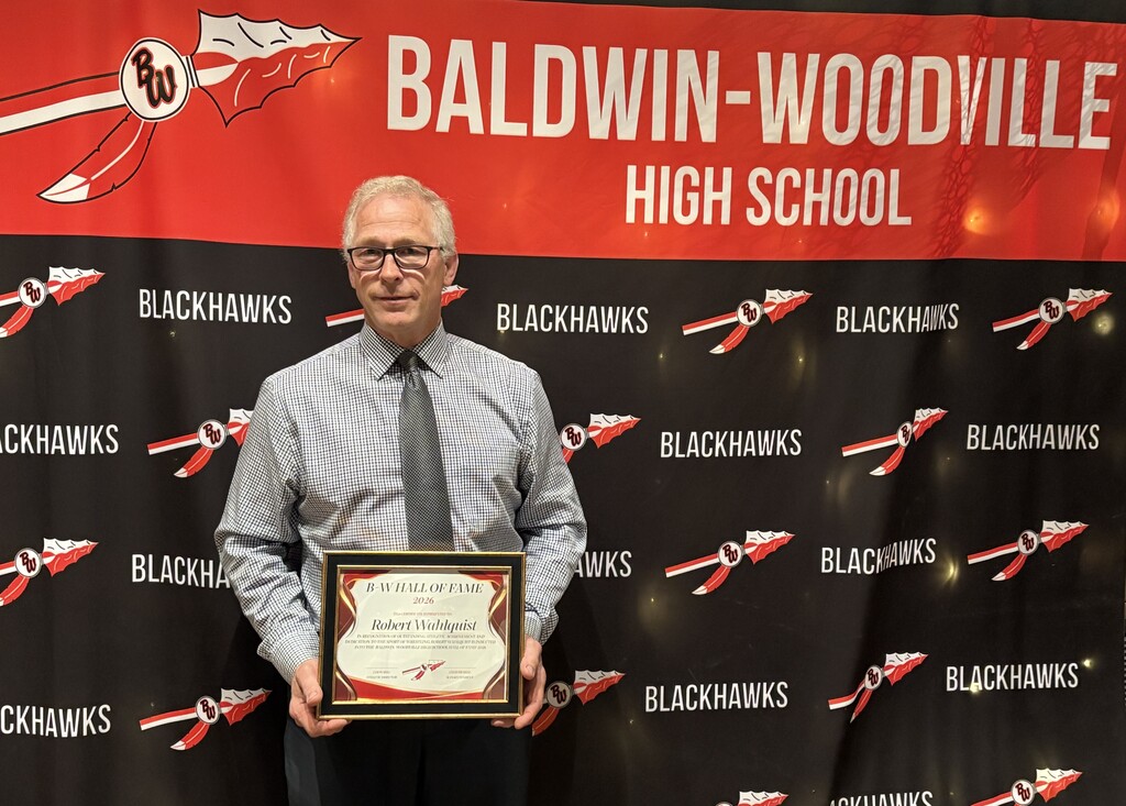 Bob Wahlquist standing in front of a red, black, and white Baldwin-Woodville High School backdrop featuring the district spear logo and "BLACKHAWKS" text. He is wearing a collared shirt and glasses, proudly holding his framed 2026 Baldwin-Woodville Hall of Fame certificate.