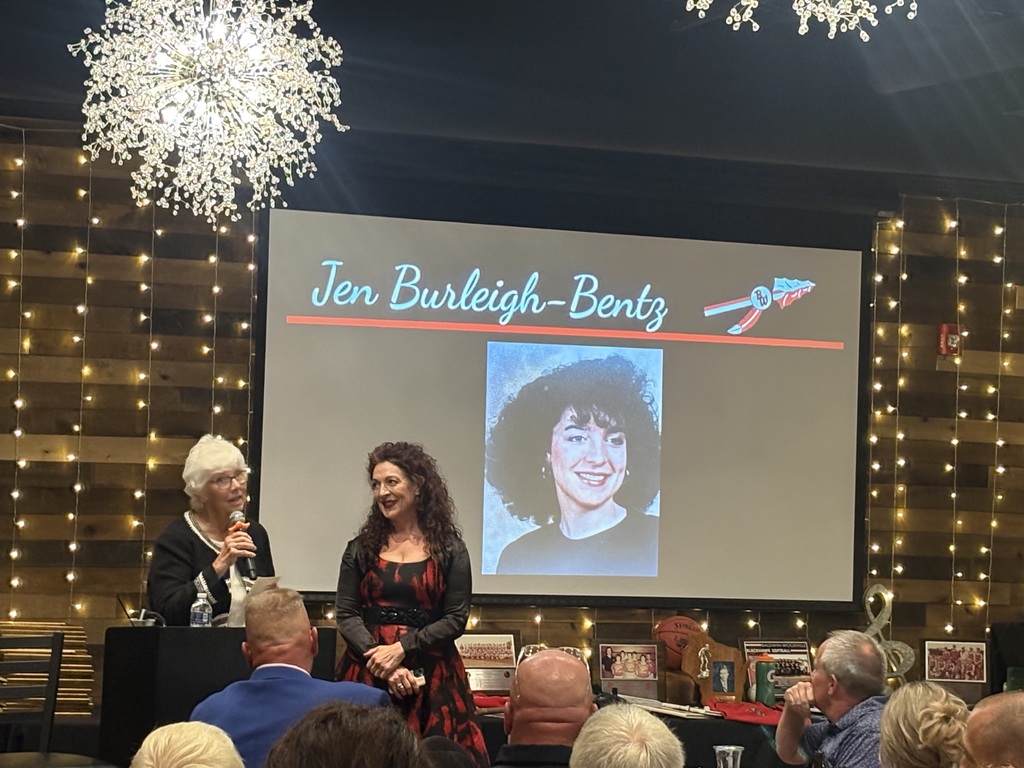 Jen Burleigh-Bentz wearing a red and black patterned dress stands next to a speaker at a podium inside a decorated venue, with a large projector screen behind them displaying her name, high school portrait, and the Baldwin-Woodville logo.