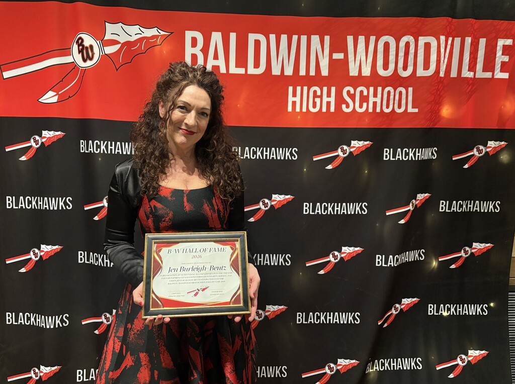 Jen Burleigh-Bentz standing in front of a red and black Baldwin-Woodville Blackhawks backdrop, smiling and holding her framed 2026 Hall of Fame induction plaque.